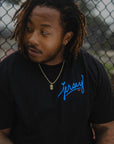 Man wearing Blue Jersey Love Tee in Black by chain link fence. Wakefield District streetwear fit showing clear blue Jersey graphic from the Contant Avenue collection.