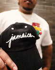 Close-up of a male model holding the Black Jamaica Signature Cord Hat toward the camera, showcasing the crisp 3D white embroidery and the structured six-panel streetwear fit.