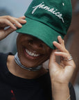 Close-up of a smiling model wearing the Jasper Green Jamaica Signature Cord Hat. The shot highlights the white 3D "jamaica" cursive embroidery and the premium fine-wale corduroy texture.