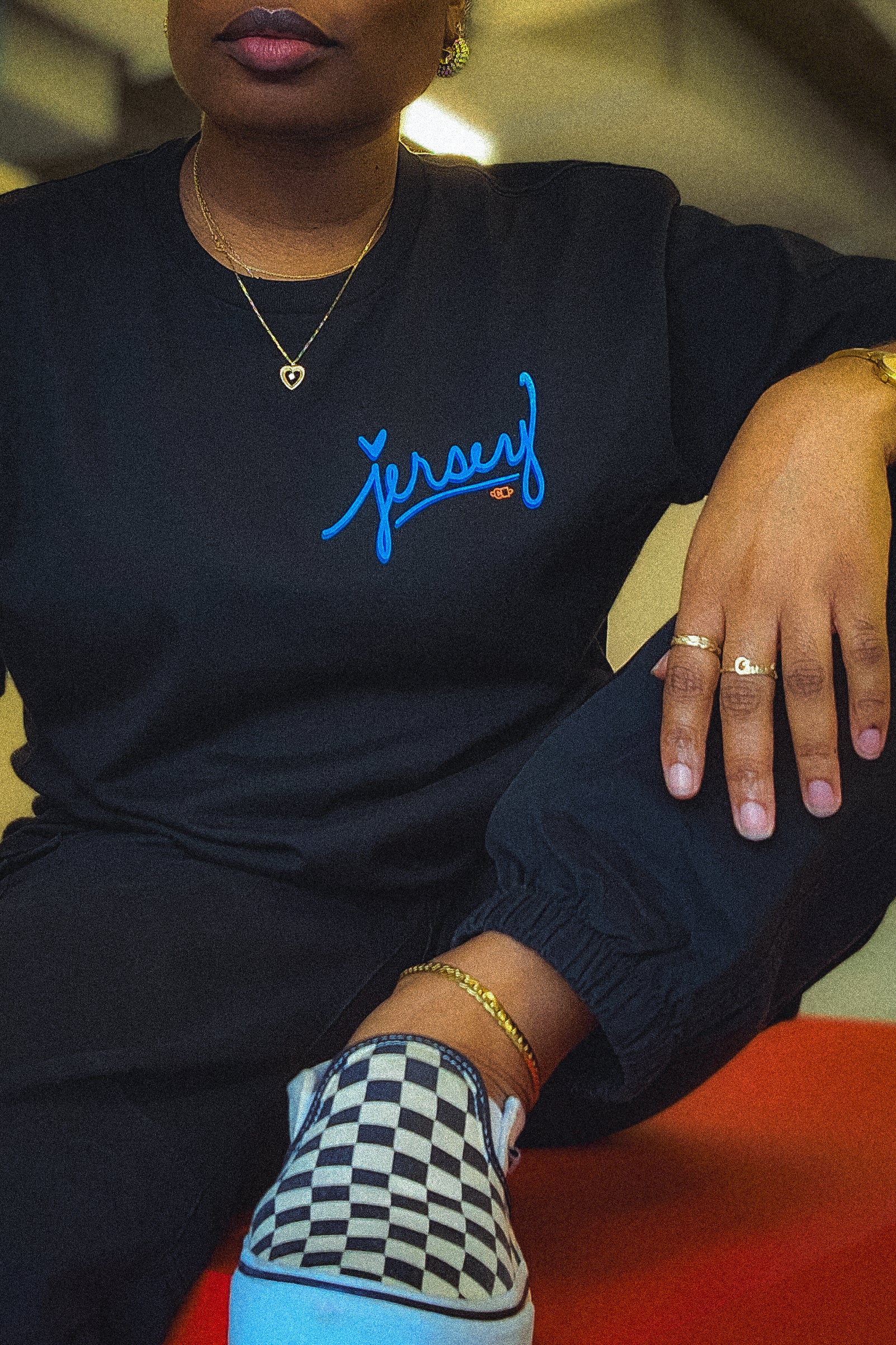 Woman wearing Blue Jersey Love Tee in Black sitting on a large red box. Wakefield District streetwear fit showing clear blue Jersey graphic from the Contant Avenue collection.
