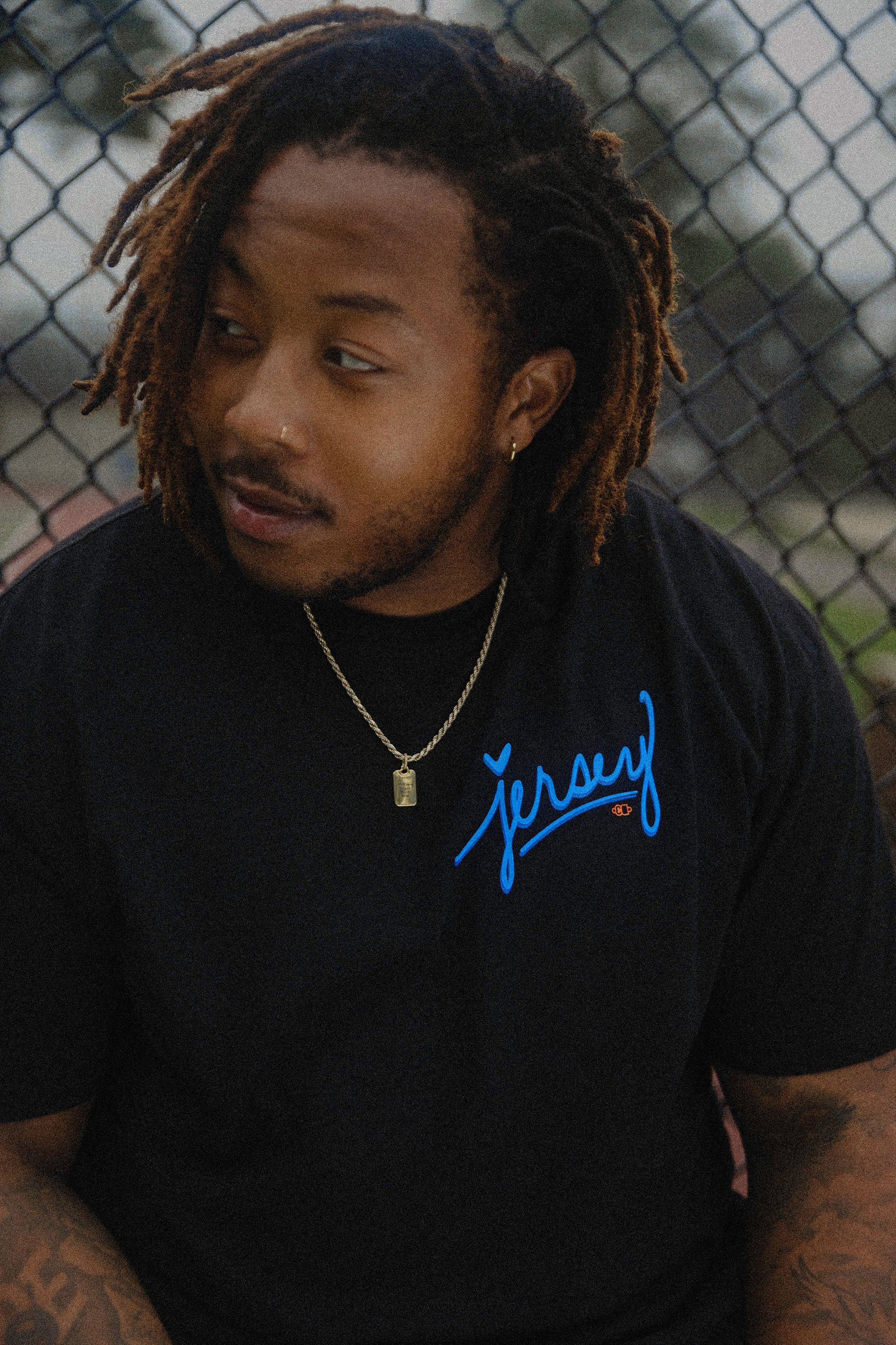 Man wearing Blue Jersey Love Tee in Black by chain link fence. Wakefield District streetwear fit showing clear blue Jersey graphic from the Contant Avenue collection.