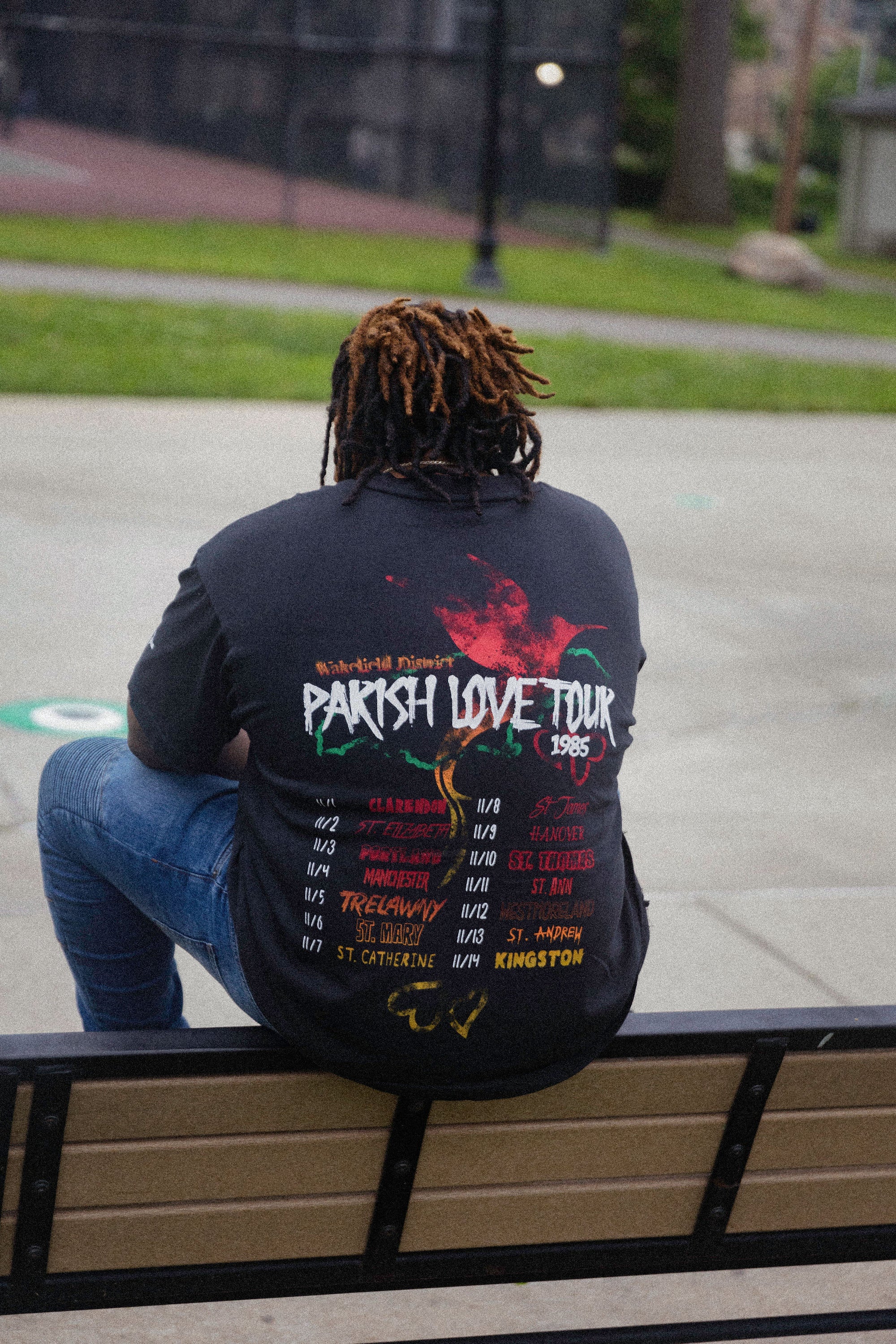man sitting on park bench wearing black parish love tour t-shirt outdoors casual streetwear