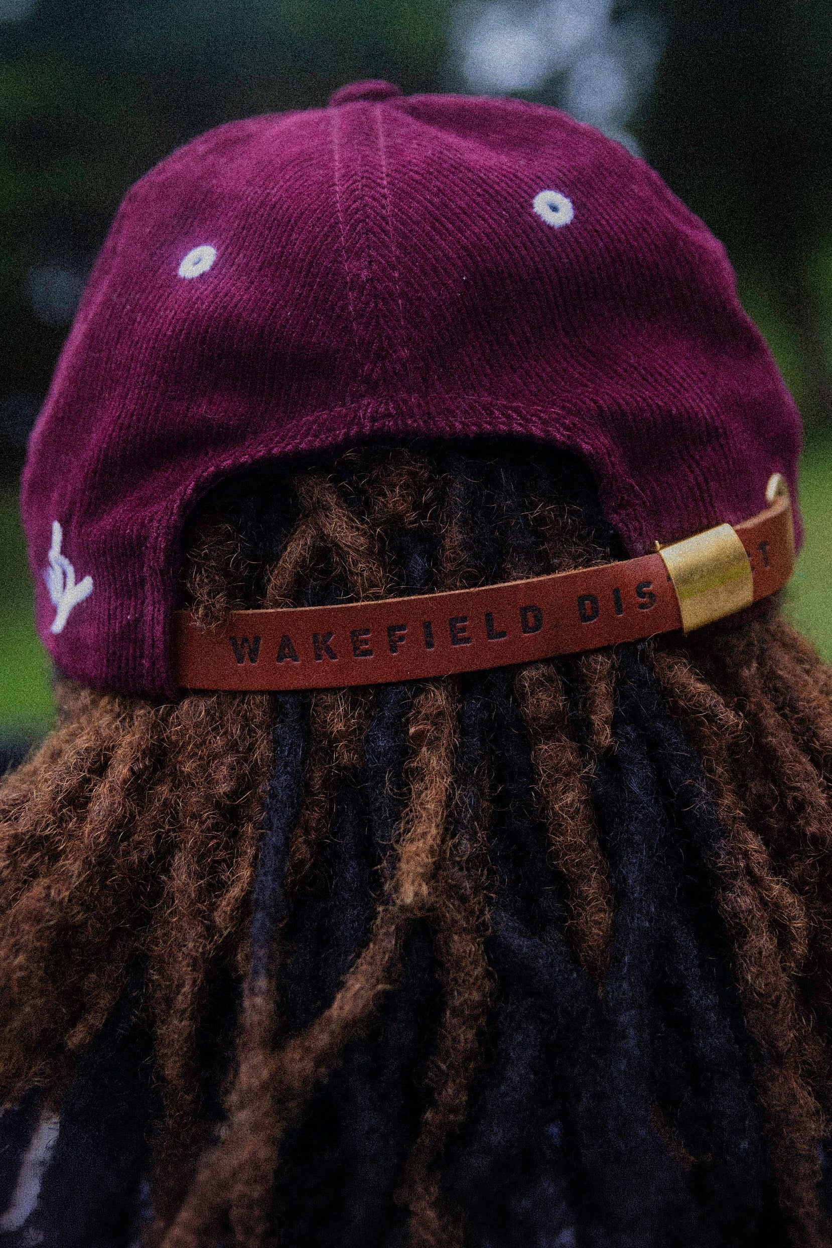 Vertical 2:3 detail shot of the back of the Garnet Red corduroy hat, featuring the adjustable hazelnut leather strap with debossed Wakefield District branding and a white embroidered logo on the side.
