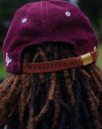 Vertical 2:3 detail shot of the back of the Garnet Red corduroy hat, featuring the adjustable hazelnut leather strap with debossed Wakefield District branding and a white embroidered logo on the side.