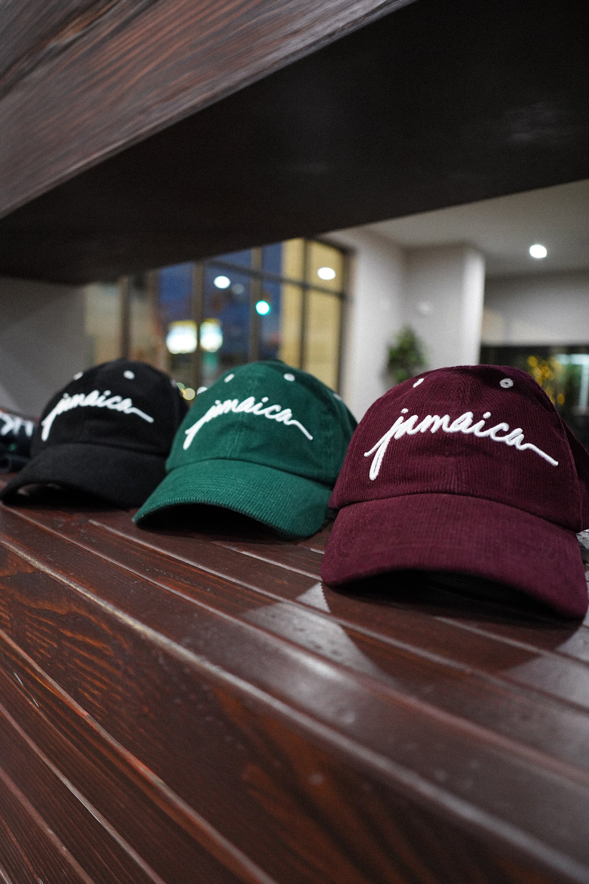 Three Wakefield District Jamaica Corduroy Hats in Black, Jasper Green, and Garnet Red displayed on a wooden ledge. The editorial film-grain style showcases the full color range of the collection.
