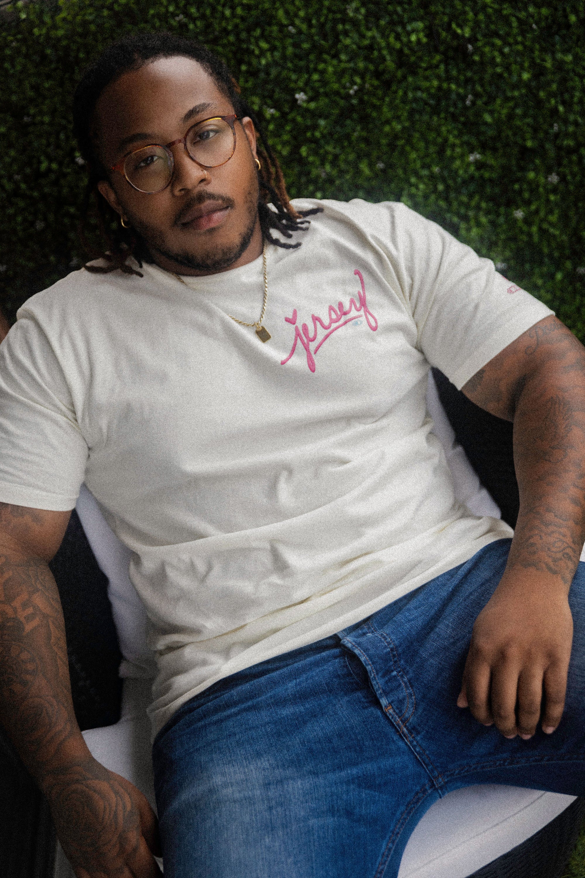A man sitting outdoors wearing a cream Wakefield District t-shirt with pink "Jersey" script across the chest, styled with blue jeans and a gold chain. Contant Avenue editorial streetwear photography.