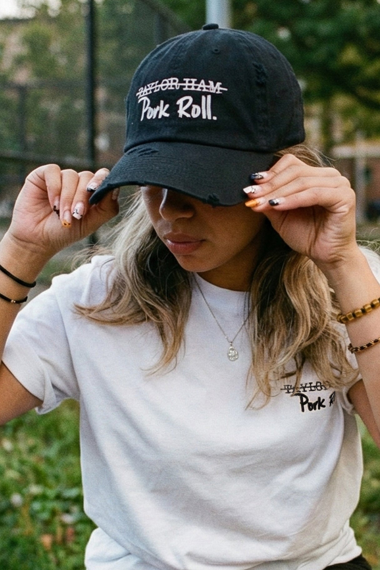 Candid film-grain photograph of a woman in an urban park, adjusting her black 'Pork Roll / Taylor Ham.' distressed hat. She is also wearing a white 'Taylor Pork Roll' t-shirt from the Wakefield District Contant Avenue collection. The background shows a chain-link fence and trees under diffused light.