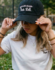 Candid film-grain photograph of a woman in an urban park, adjusting her black 'Pork Roll / Taylor Ham.' distressed hat. She is also wearing a white 'Taylor Pork Roll' t-shirt from the Wakefield District Contant Avenue collection. The background shows a chain-link fence and trees under diffused light.
