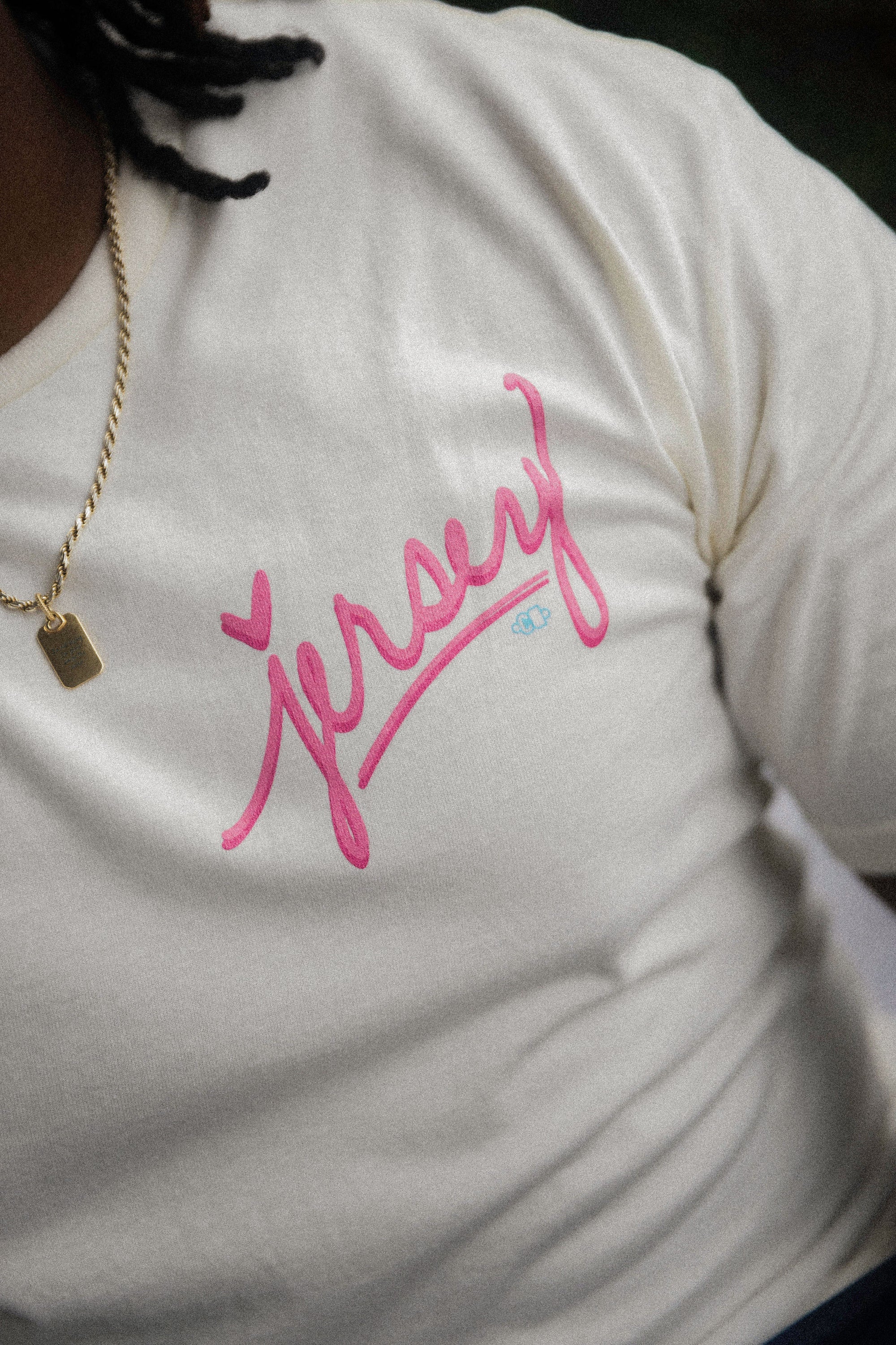 Close-up of a cream Wakefield District t-shirt featuring a prominent pink "Jersey" script graphic with a heart accent on the chest, paired with a gold chain. Contant Avenue collection detail shot.