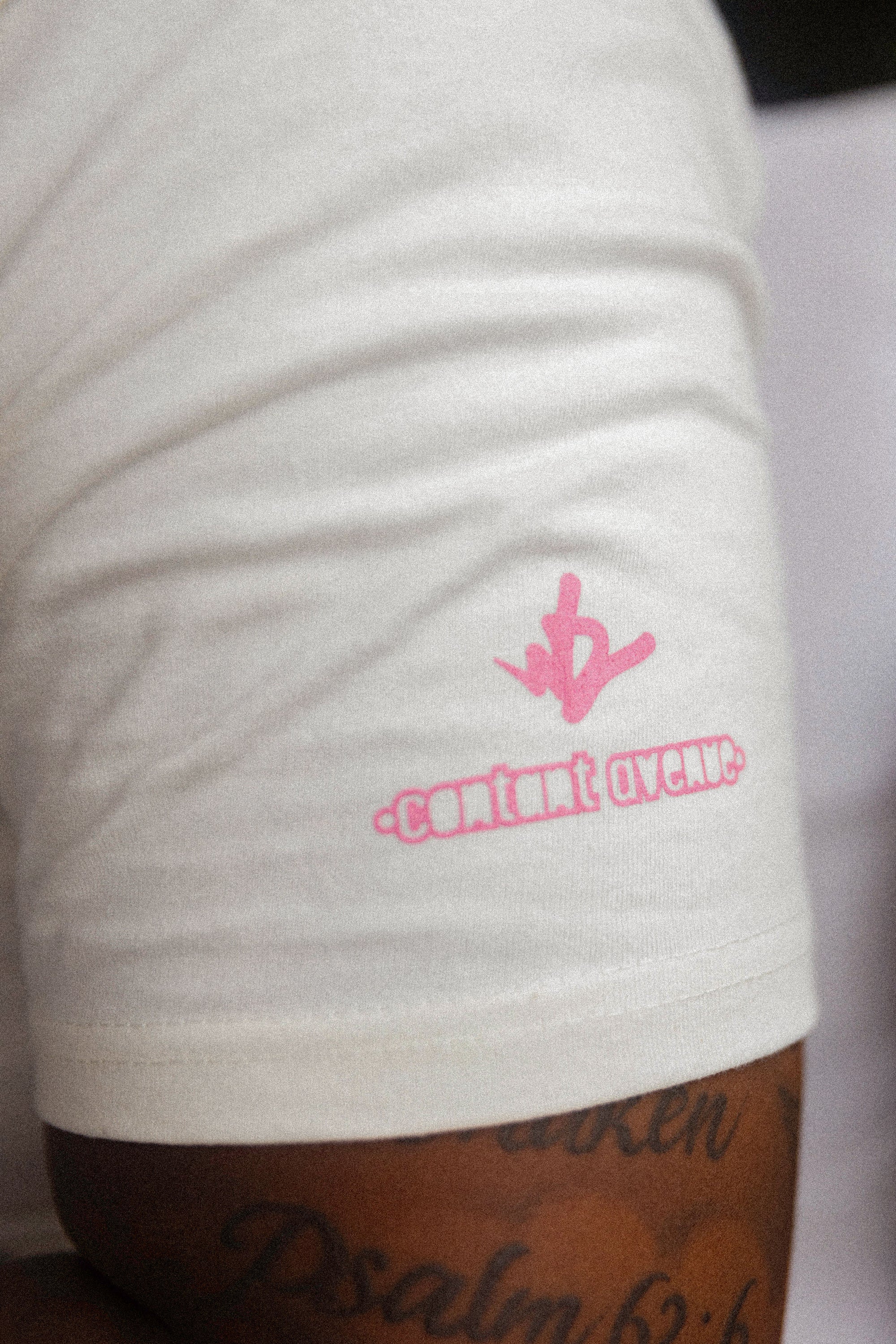 Close-up detail of the sleeve on a cream Wakefield District t-shirt, showing pink WD and Contant Avenue logos. New Jersey streetwear aesthetic.