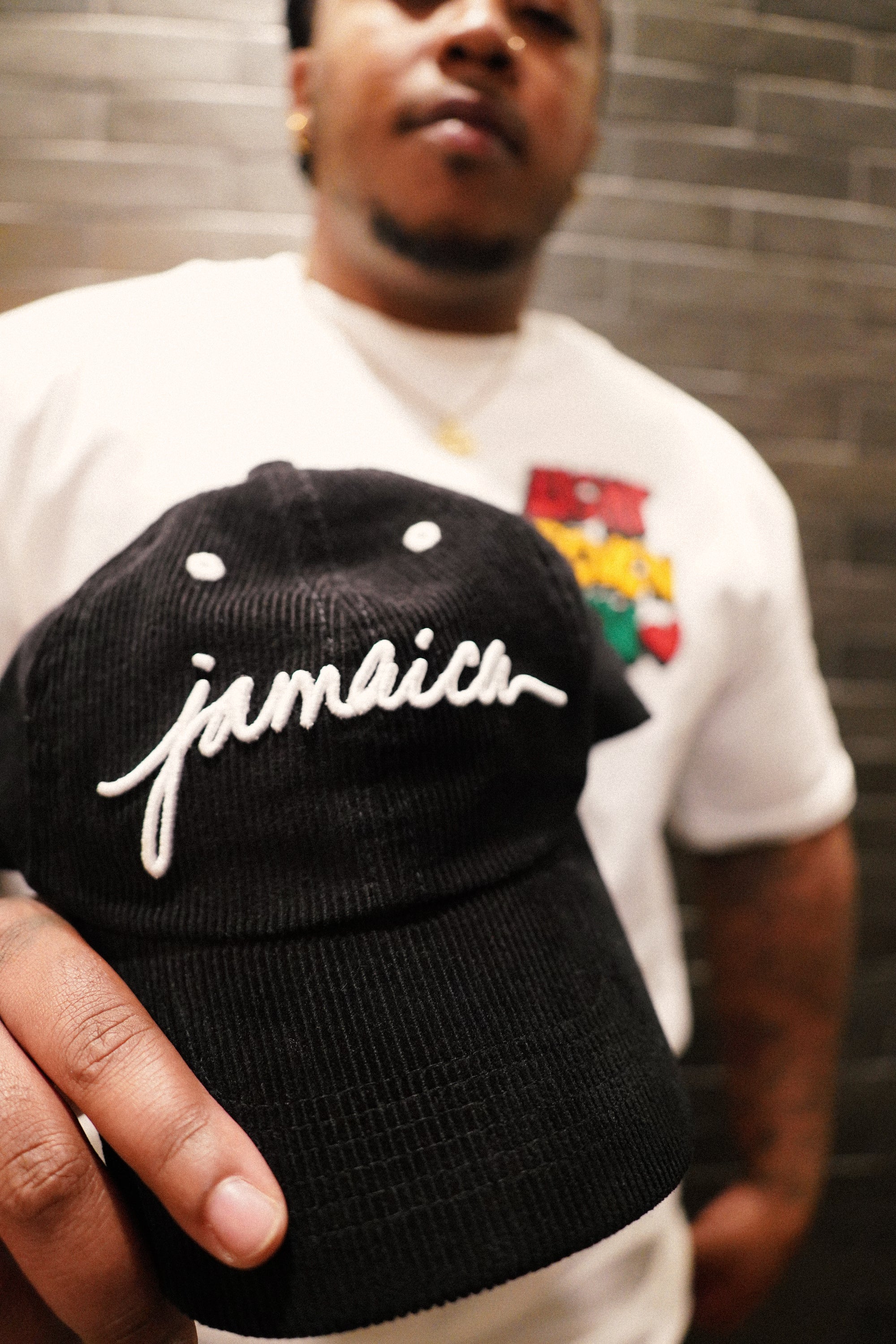 Close-up of a male model holding the Black Jamaica Signature Cord Hat toward the camera, showcasing the crisp 3D white embroidery and the structured six-panel streetwear fit.