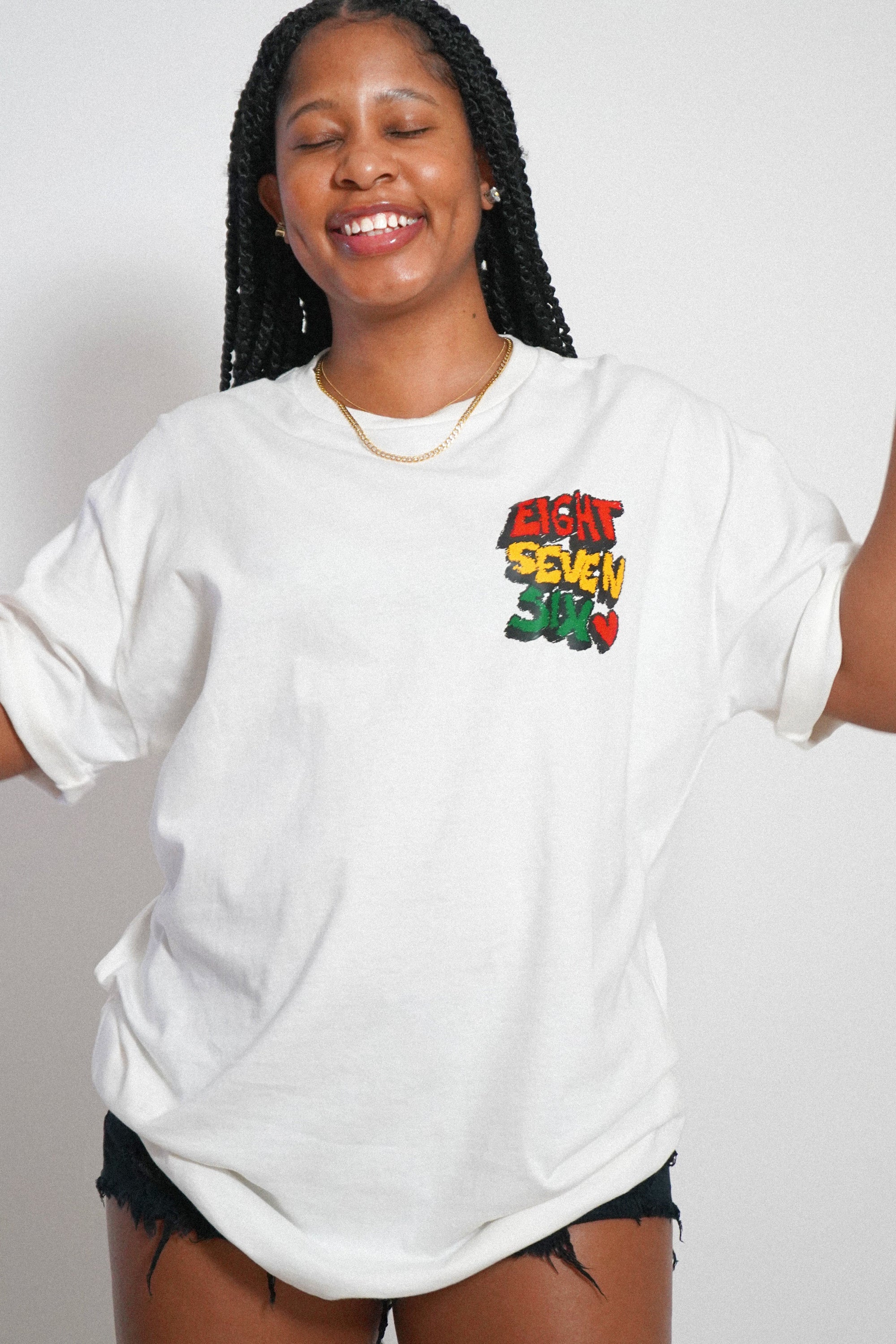 Female model with braided hair smiling and wearing the white Eight Seven Six Scribble Tee, showcasing the relaxed fit and the custom 876 chest graphic. Studio photograph with a cohesive warm, grained film aesthetic.