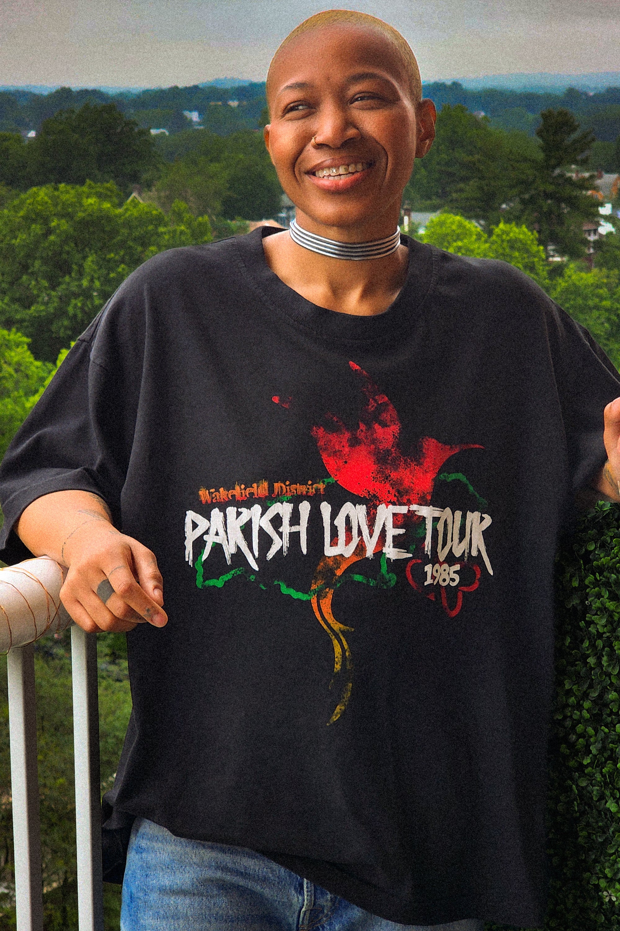 Model smiling on a balcony wearing the Wakefield District Parish Love Tour Tee in vintage black, showcasing the front distressed red, green, and yellow hummingbird graphic and the garment's relaxed, oversized streetwear fit.