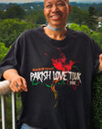 Model smiling on a balcony wearing the Wakefield District Parish Love Tour Tee in vintage black, showcasing the front distressed red, green, and yellow hummingbird graphic and the garment's relaxed, oversized streetwear fit.