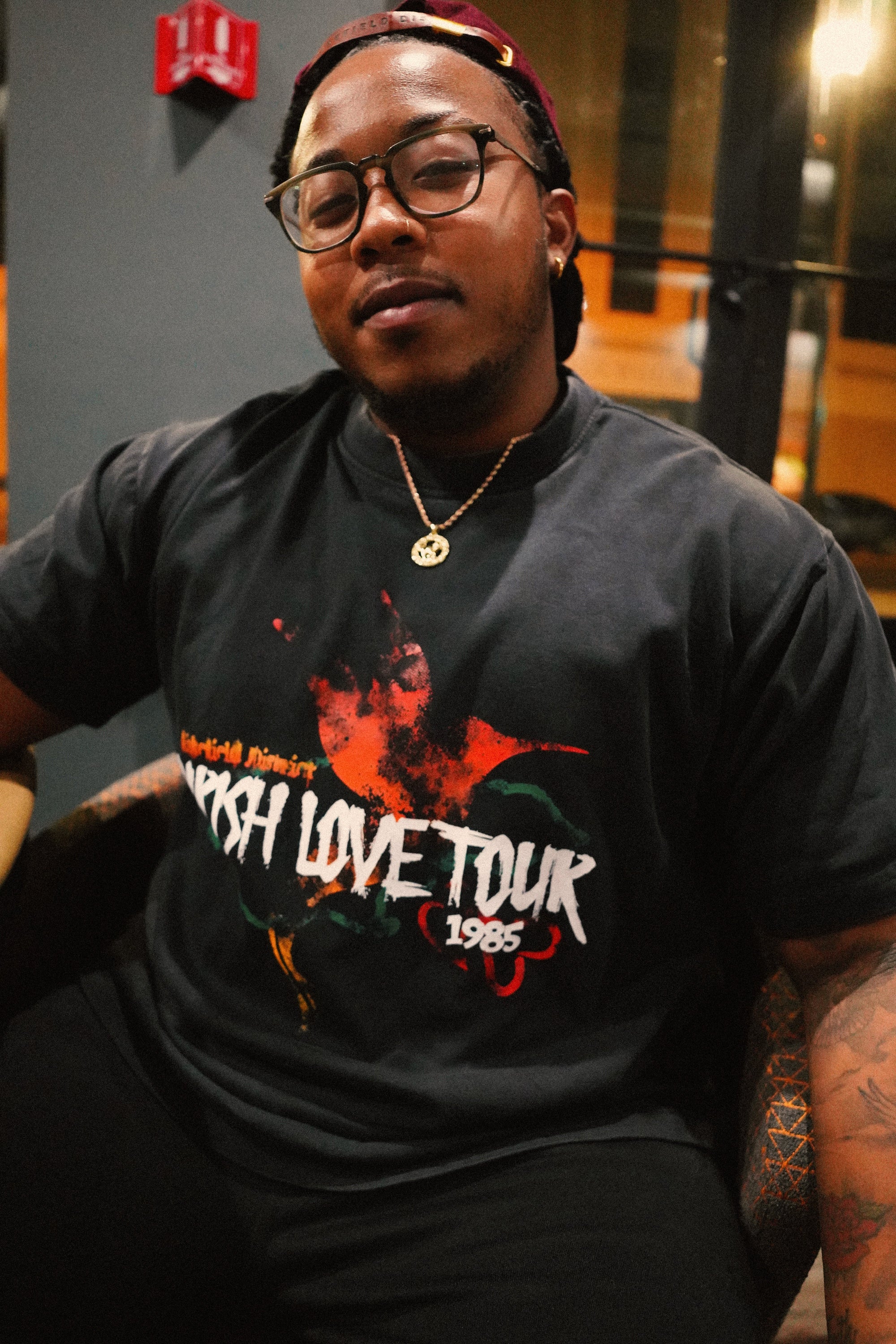 Male model wearing the Wakefield District Parish Love Tour Tee in vintage black, demonstrating the relaxed, unisex streetwear fit. He is seated indoors, styling the shirt with a burgundy hat and glasses, showcasing the distressed red and green front hummingbird graphic.