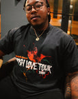Male model wearing the Wakefield District Parish Love Tour Tee in vintage black, demonstrating the relaxed, unisex streetwear fit. He is seated indoors, styling the shirt with a burgundy hat and glasses, showcasing the distressed red and green front hummingbird graphic.