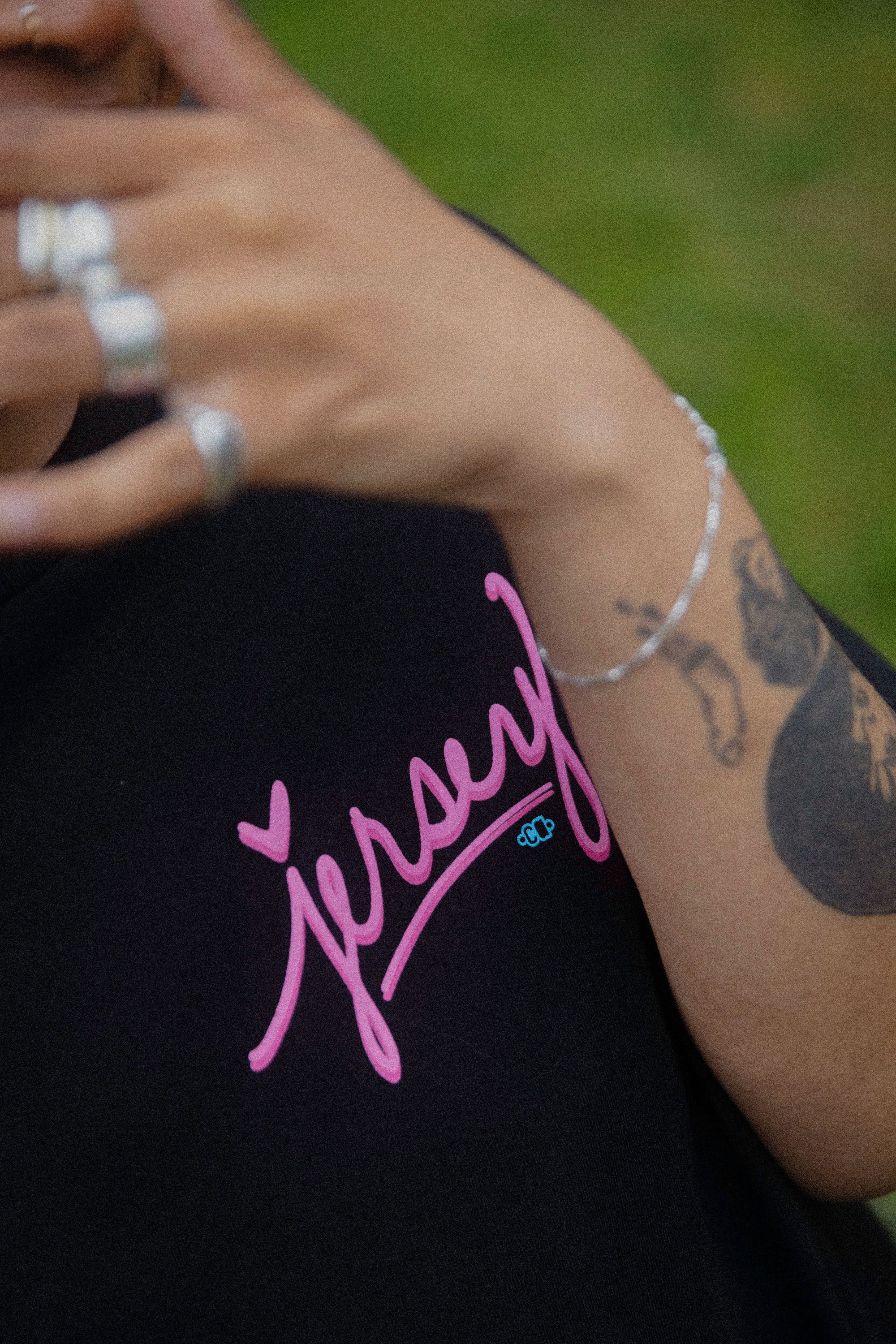 Close-up lifestyle detail of a black Wakefield District t-shirt featuring a pink cursive Jersey logo, worn by a model with arm tattoos and silver jewelry.