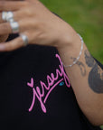 Close-up lifestyle detail of a black Wakefield District t-shirt featuring a pink cursive Jersey logo, worn by a model with arm tattoos and silver jewelry.